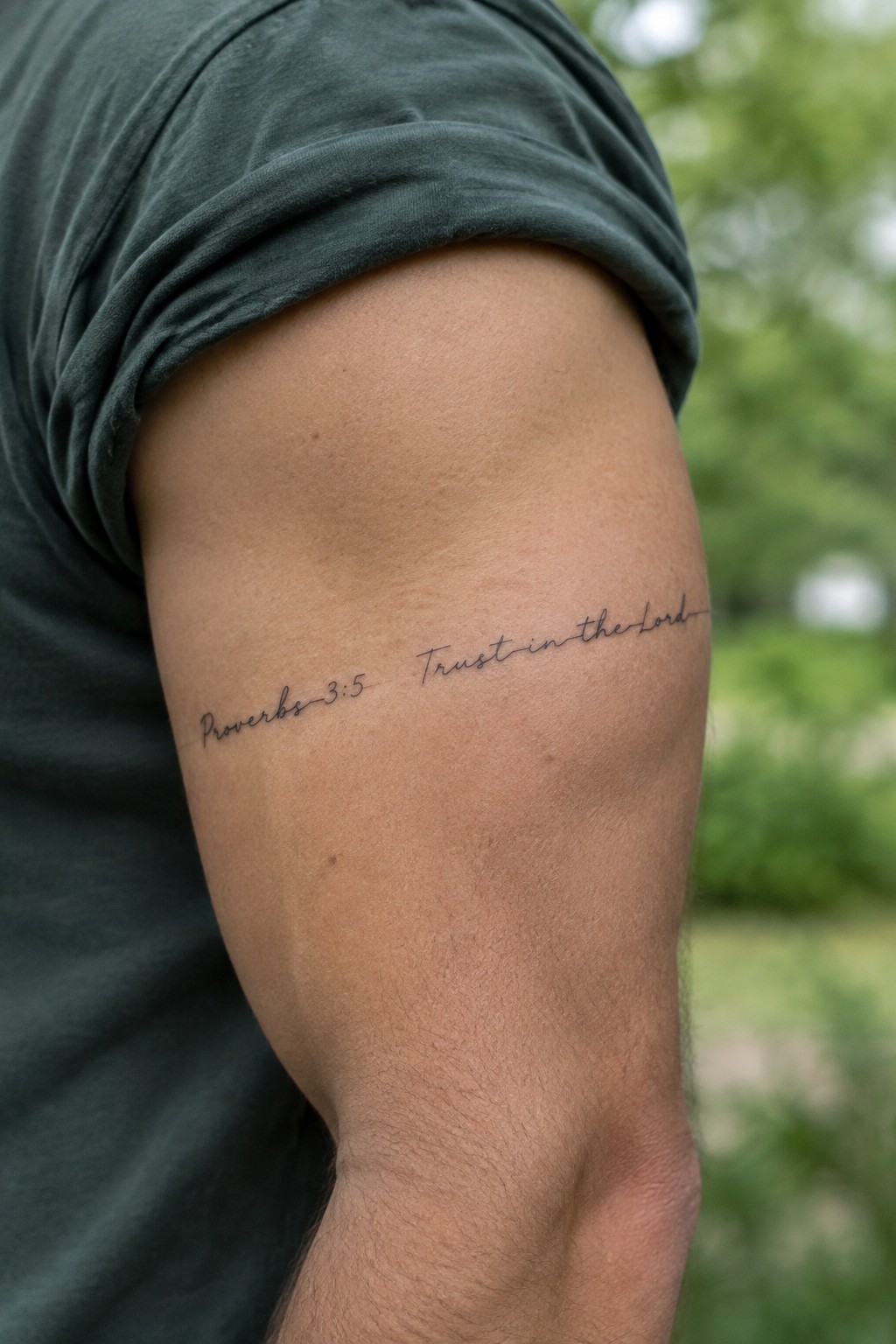 Close up of a thin cursive tattoo on a man's upper arm showing the text Proverbs 3:5 Trust in the Lord