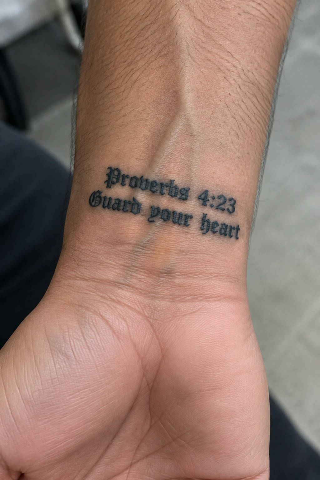 Close up of a Proverbs 4:23 Guard your heart tattoo in gothic font on the inner wrist of a man with medium skin tone.