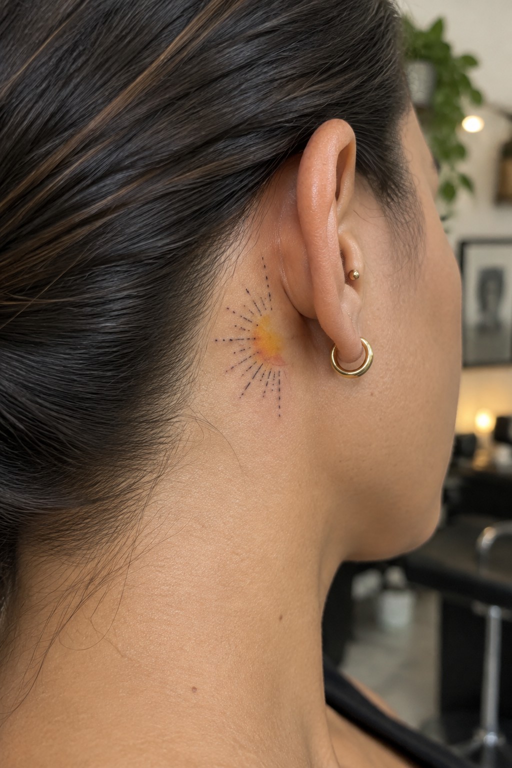 Sunburst tattoo with yellow-orange center and black rays behind ear with gold hoop earring