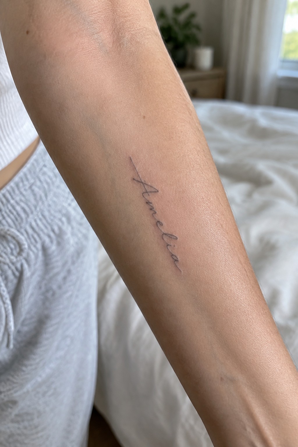 Cursive black 'Amelia' tattoo on a forearm beside white bedding and a nightstand