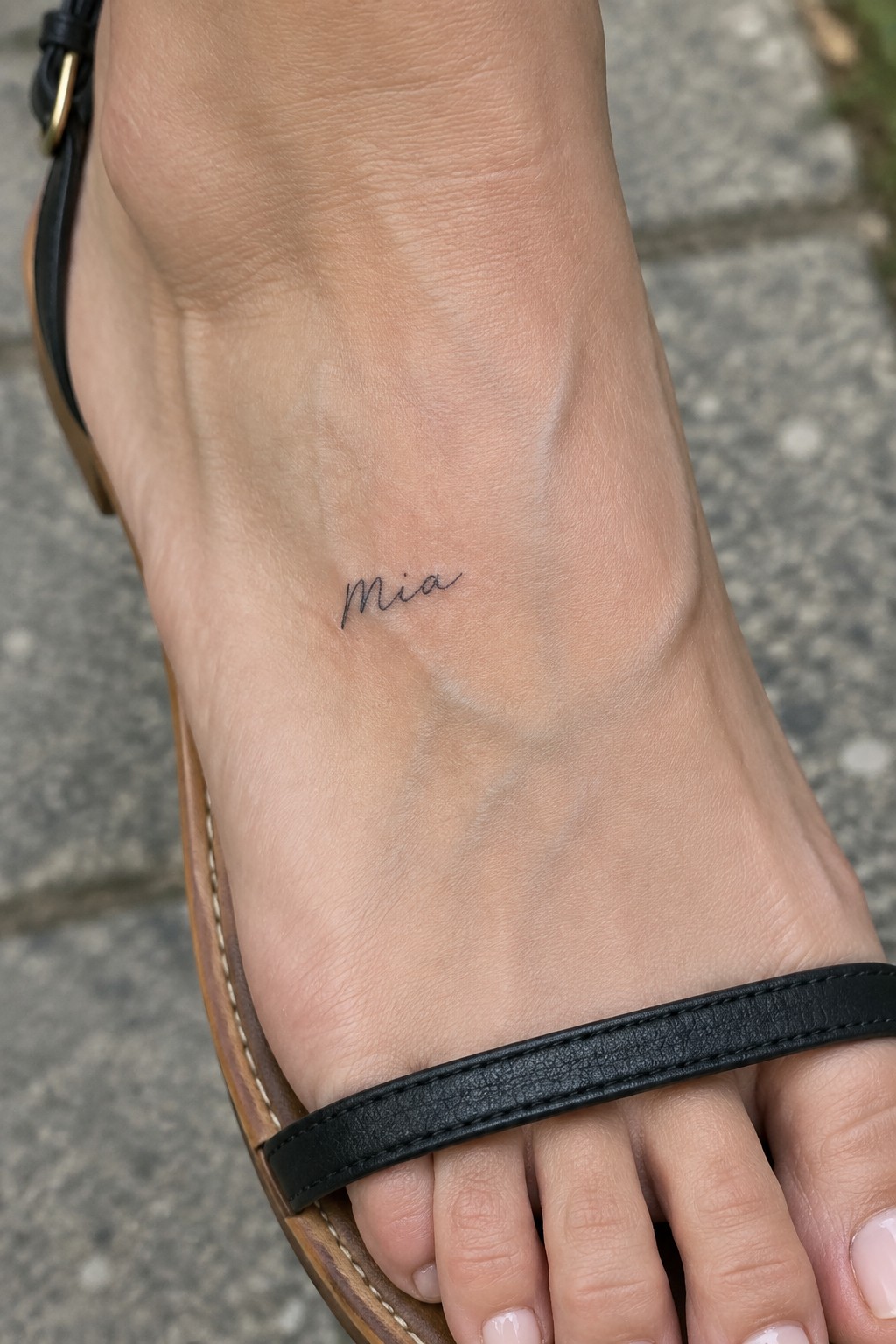 Foot with cursive 'Mia' tattoo wearing black leather sandal on pavement.