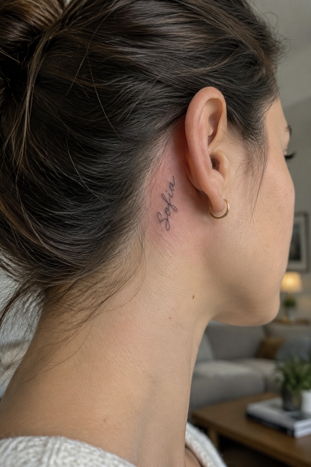 Woman with Sofia tattoo behind ear, brown hair in bun, gold hoop earring.