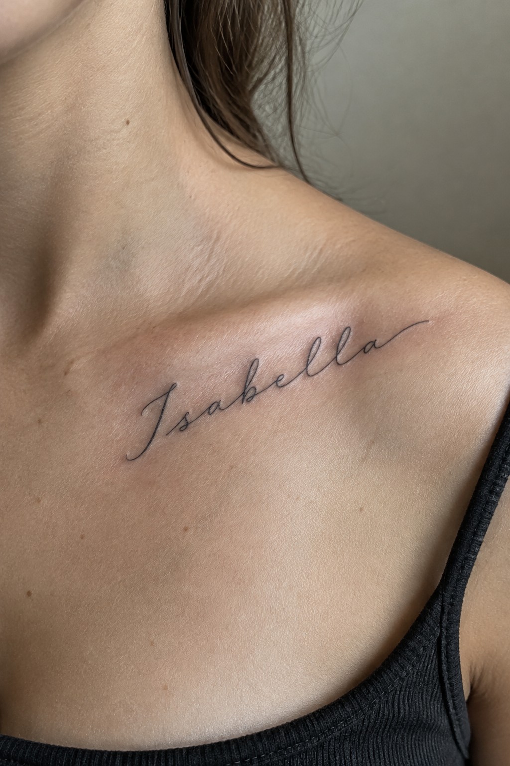 Fine line handwritten tattoo of the name Isabella in cursive script placed on a woman's collarbone area.