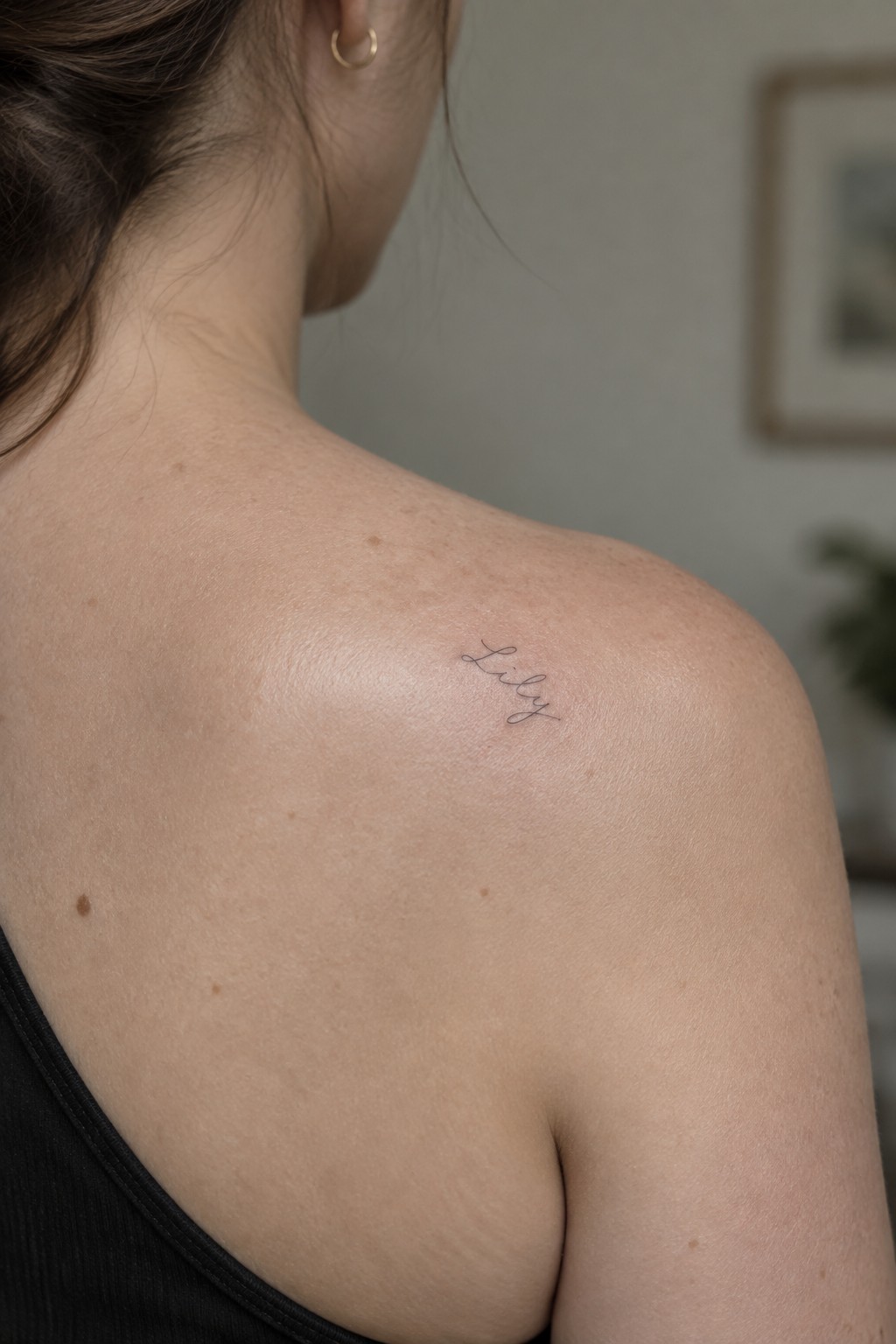 Woman's freckled shoulder with cursive 'Lily' tattoo, black tank top, gold earring.