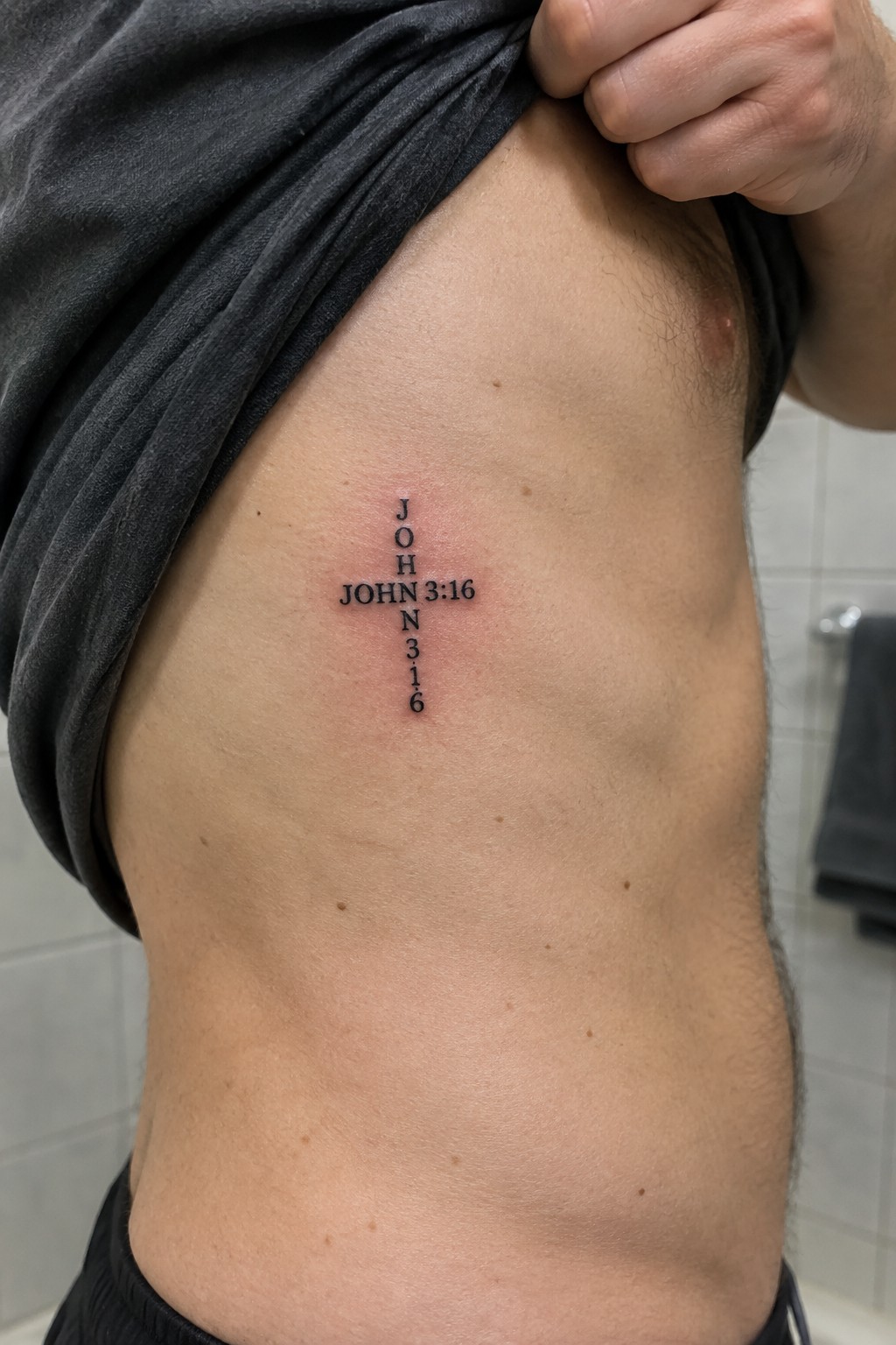 Small black cross tattoo with John 3:16 text integrated vertically on a man's side torso.