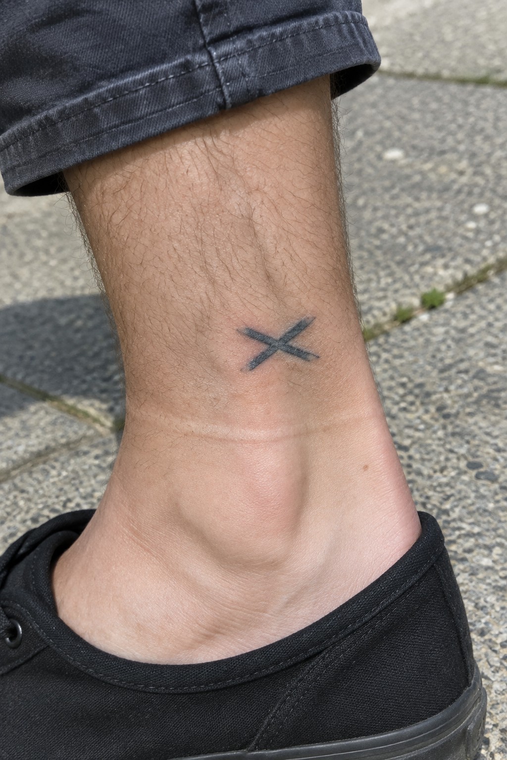 Black X tattoo on ankle with dark rolled-up jeans and black sneaker on pavement.