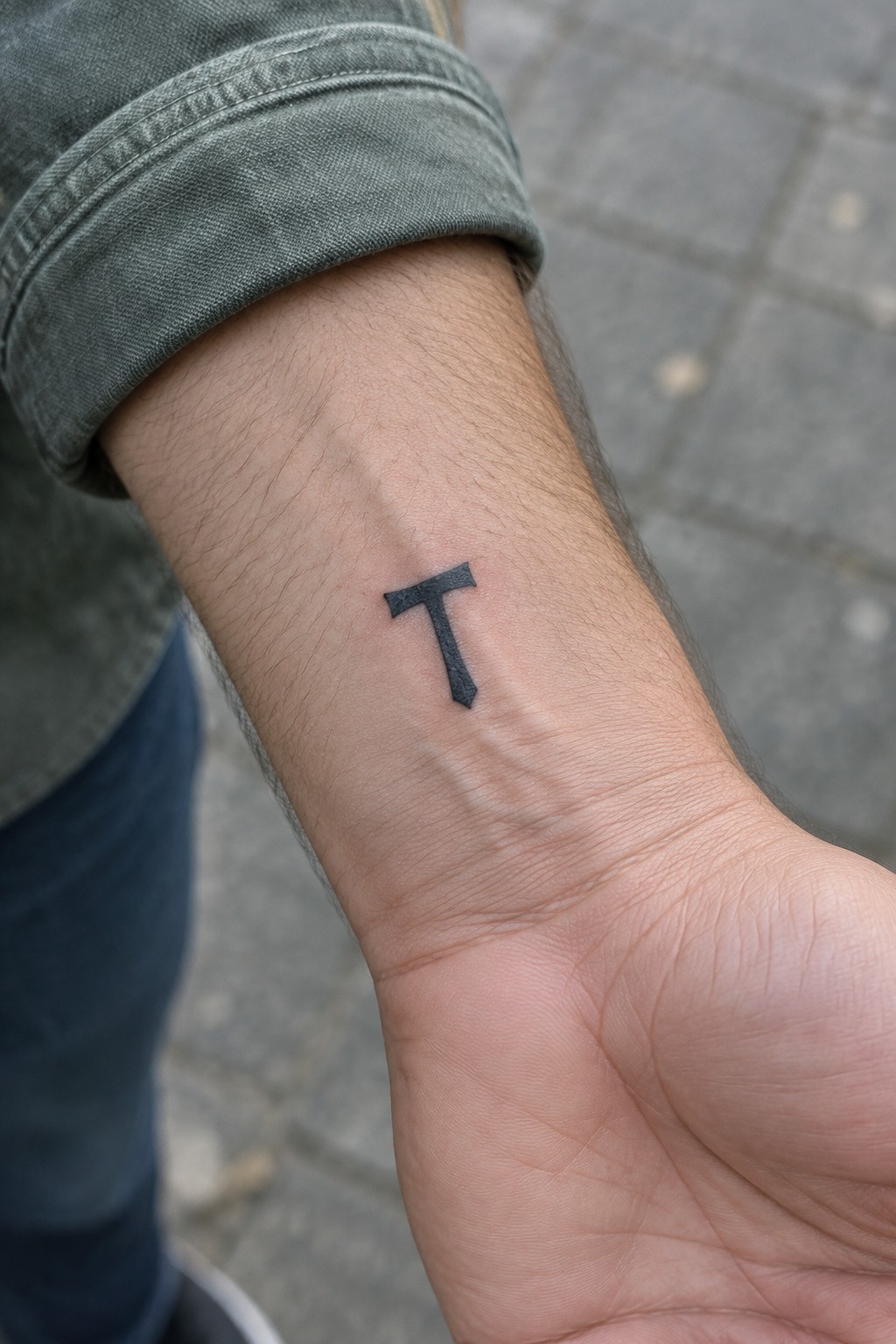 Black T tattoo on wrist under rolled-up green denim sleeve