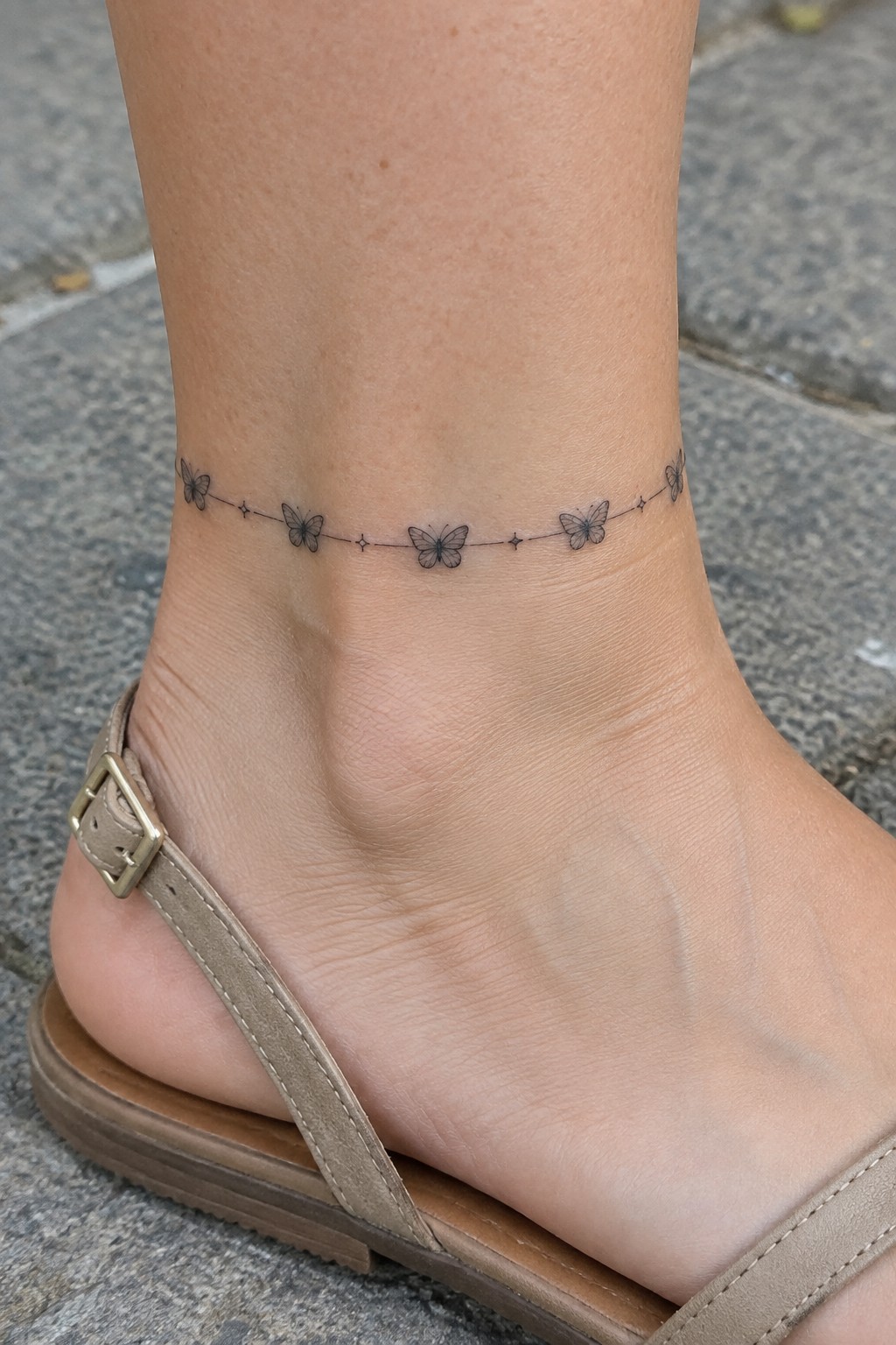 Ankle tattoo of butterflies linked by dotted line above beige sandal on stone pavement