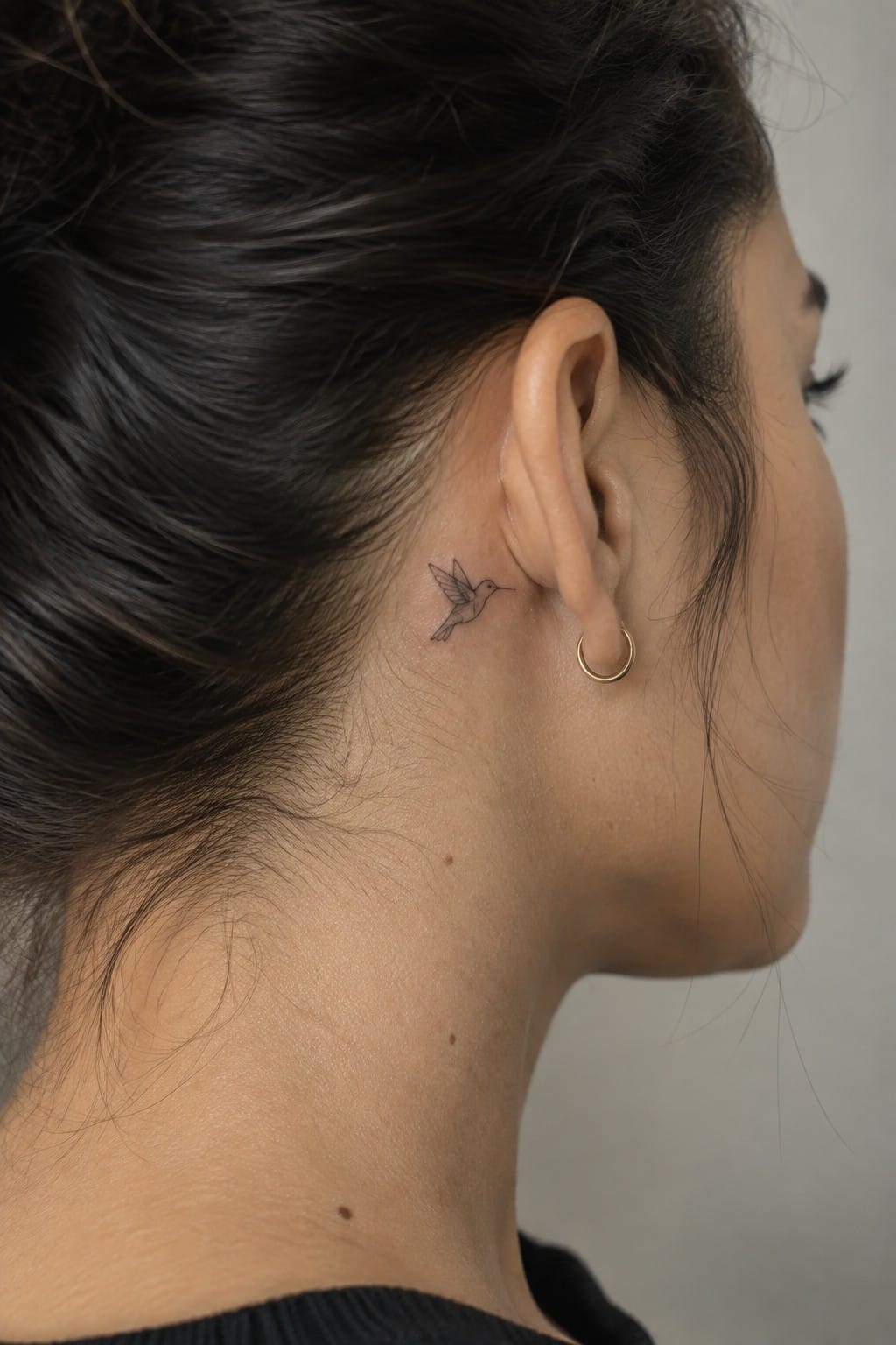 Woman with dark bun, hummingbird tattoo behind ear, gold hoop earring, black top