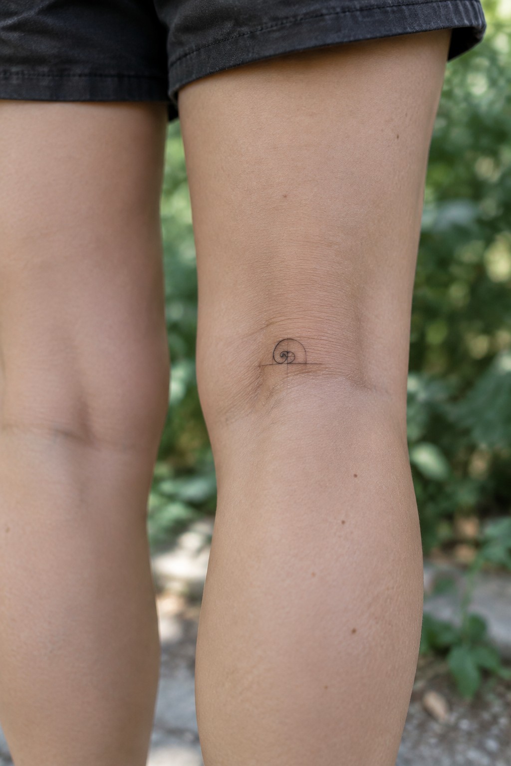 Knee with small nautilus shell tattoo, dark shorts above blurred greenery.