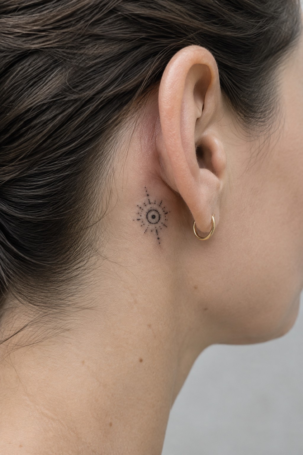 Close-up of ear with black sunburst tattoo and small gold hoop earring.