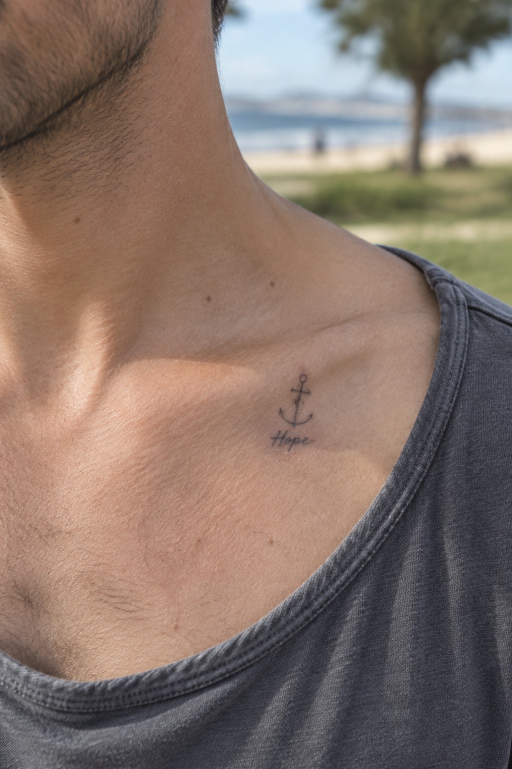 Man's collarbone with small anchor Hope tattoo above gray t-shirt, blurred beach background