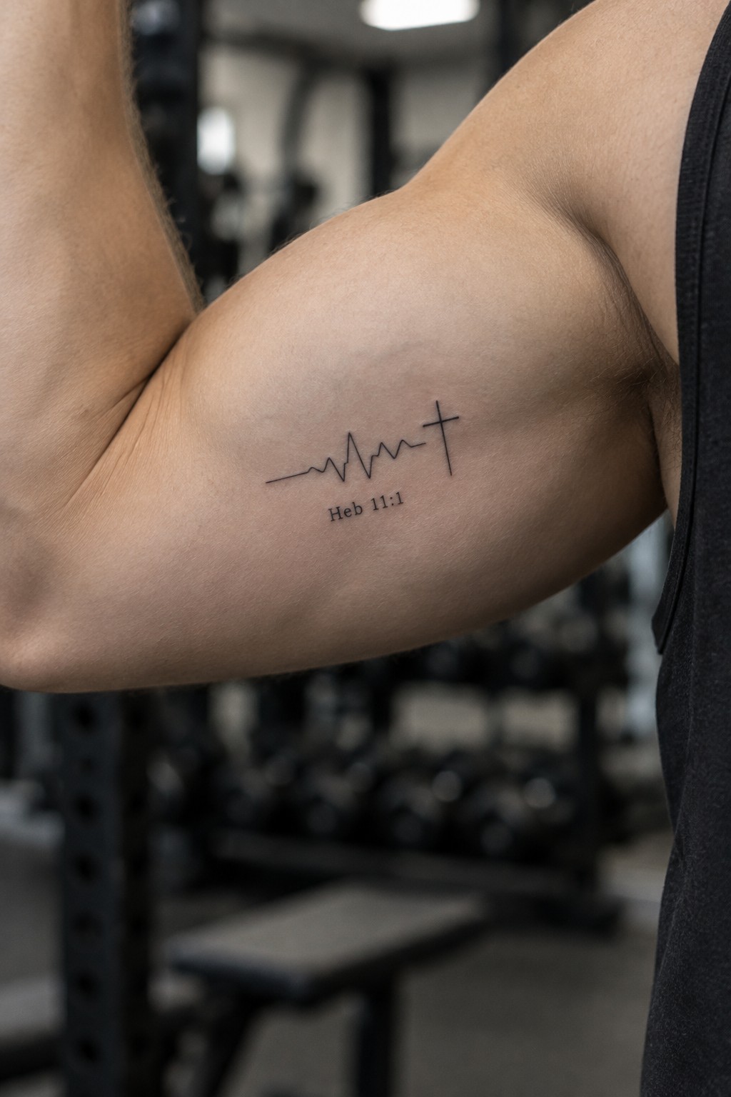 Muscular arm in black tank top with heartbeat-to-cross tattoo and Heb 11:1 in gym.