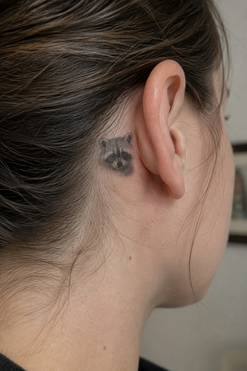 Small grayscale raccoon face tattoo positioned behind the ear on the side of the head.