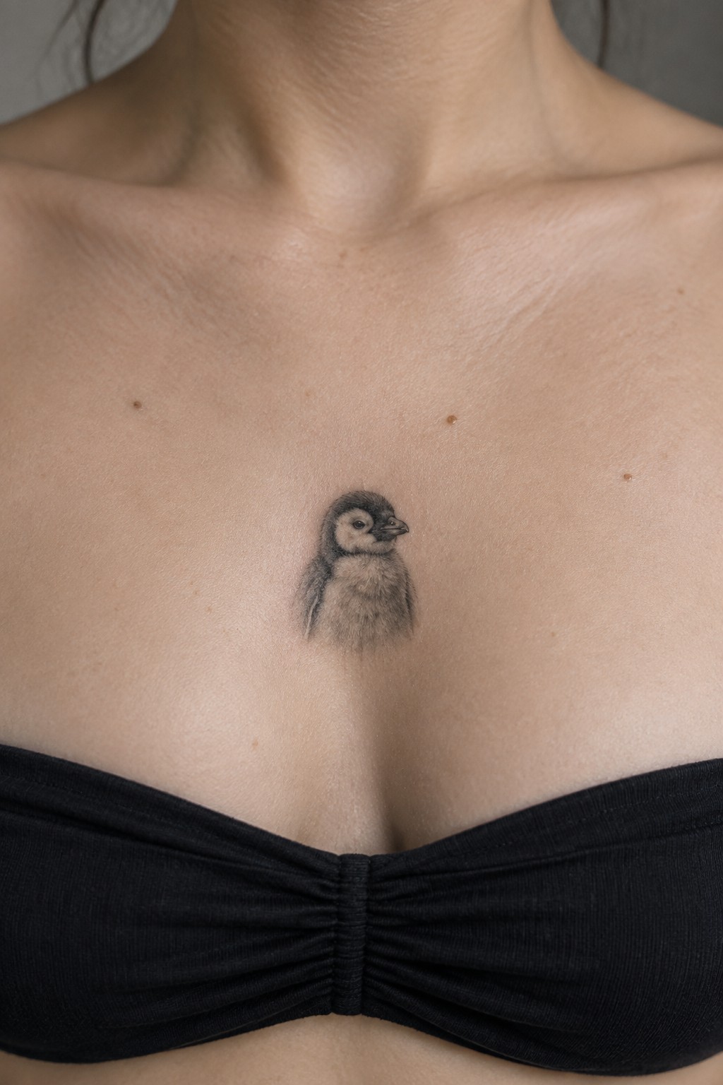 Realistic black-and-gray penguin chick tattoo on chest above black bandeau top