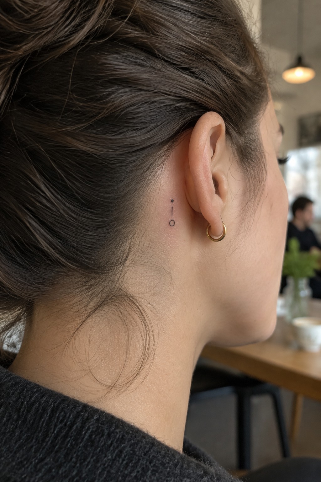 Dark brown hair in bun with small black tattoo behind ear and gold hoop earring