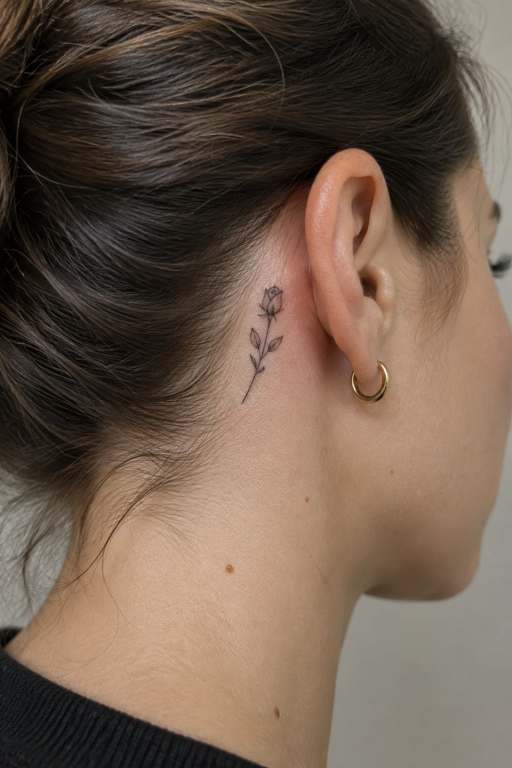 Fine line rose tattoo with stem and leaves placed behind a woman's ear