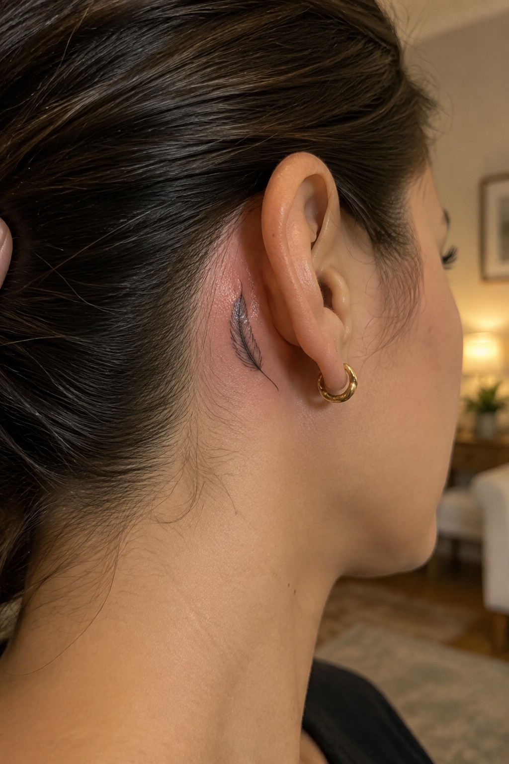 Profile view of ear with black feather tattoo and gold hoop earring on dark hair.