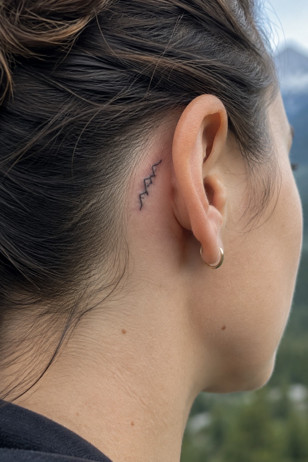 Dark brown hair in bun, small mountain tattoo behind ear with gold hoop earring