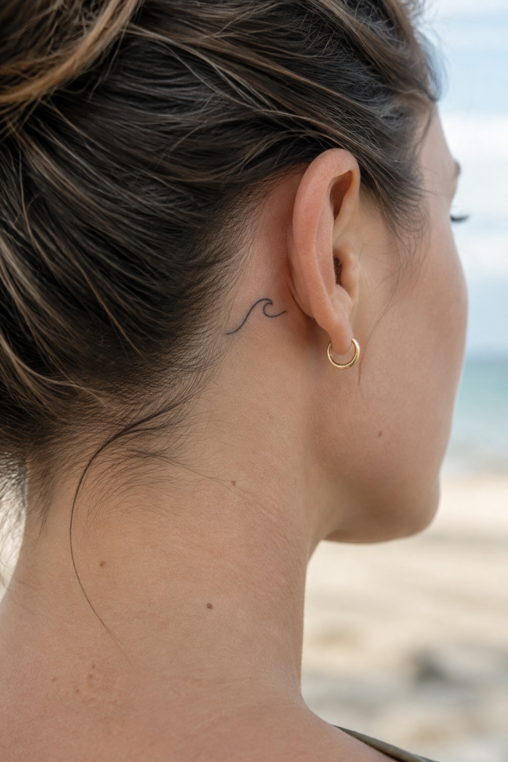 Small fine line wave tattoo placed behind the ear on a woman with pulled back hair, shown from the side against a beach background.