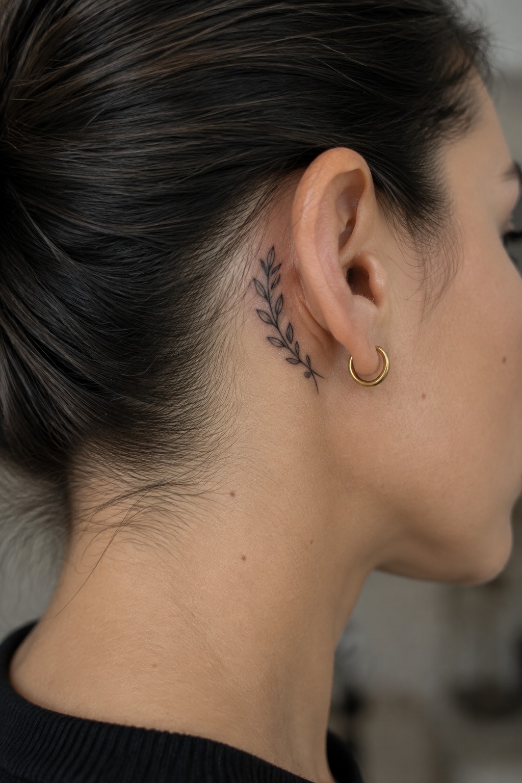 Fine line laurel branch tattoo placed behind a woman's ear on the side of the head