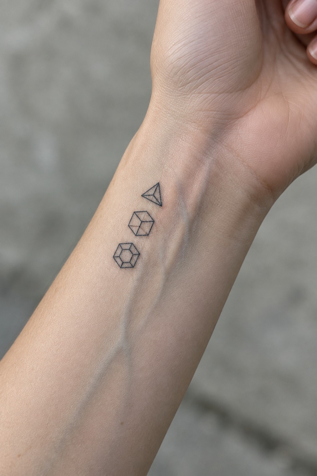 Wrist with three black line-art geometric tattoos: triangle, cube, hexagon