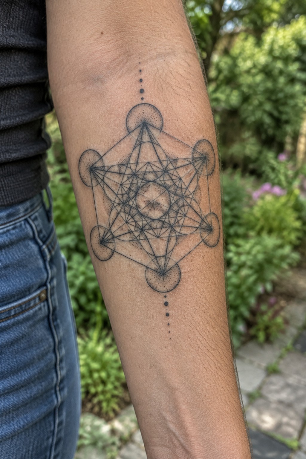 Black ink sacred geometry tattoo on a woman's inner forearm showing a detailed star pattern inside a cube like structure with circles at the points and vertical dotted lines extending from the design.
