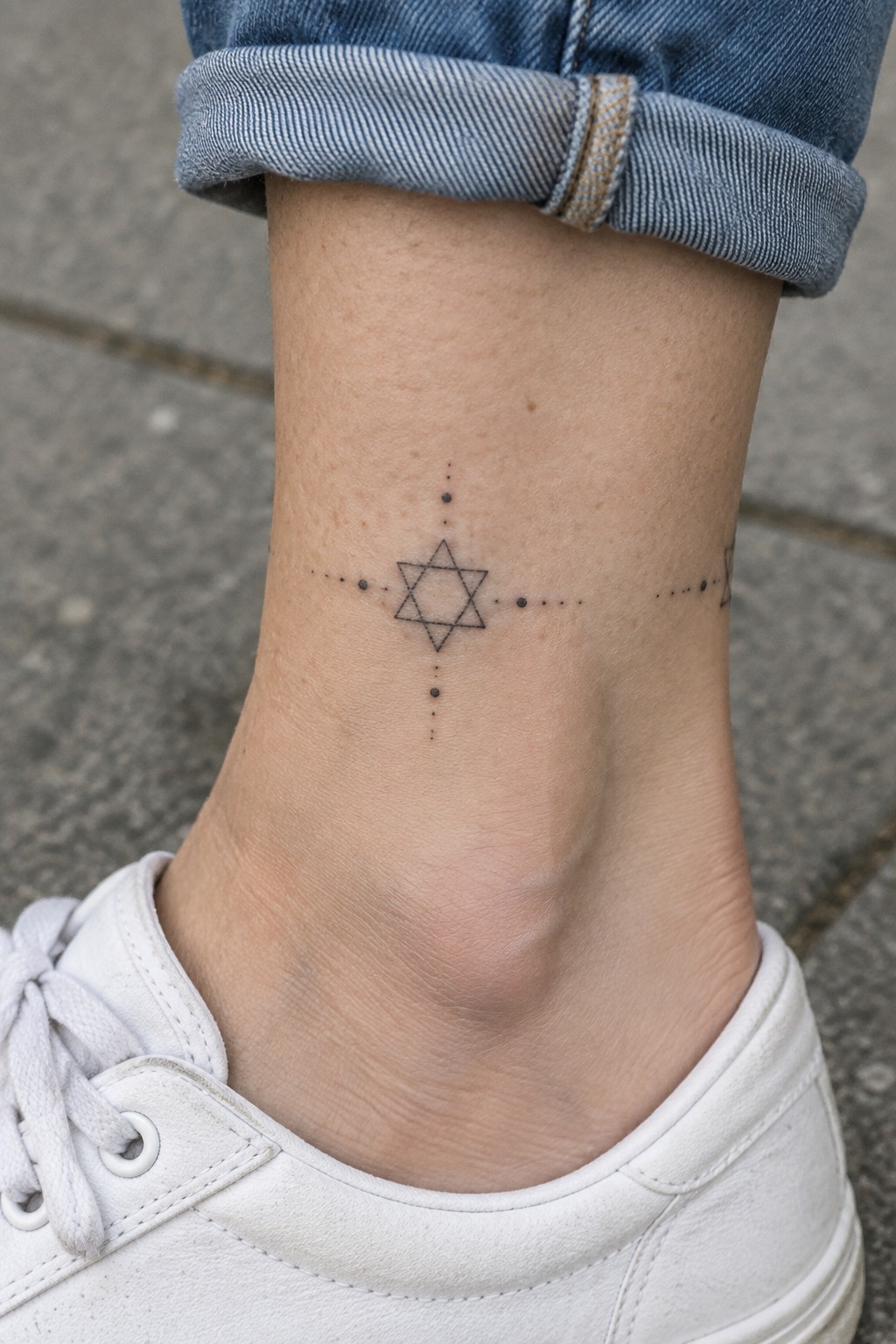 Ankle with Star of David tattoo, dotted lines, rolled blue jeans, white sneaker