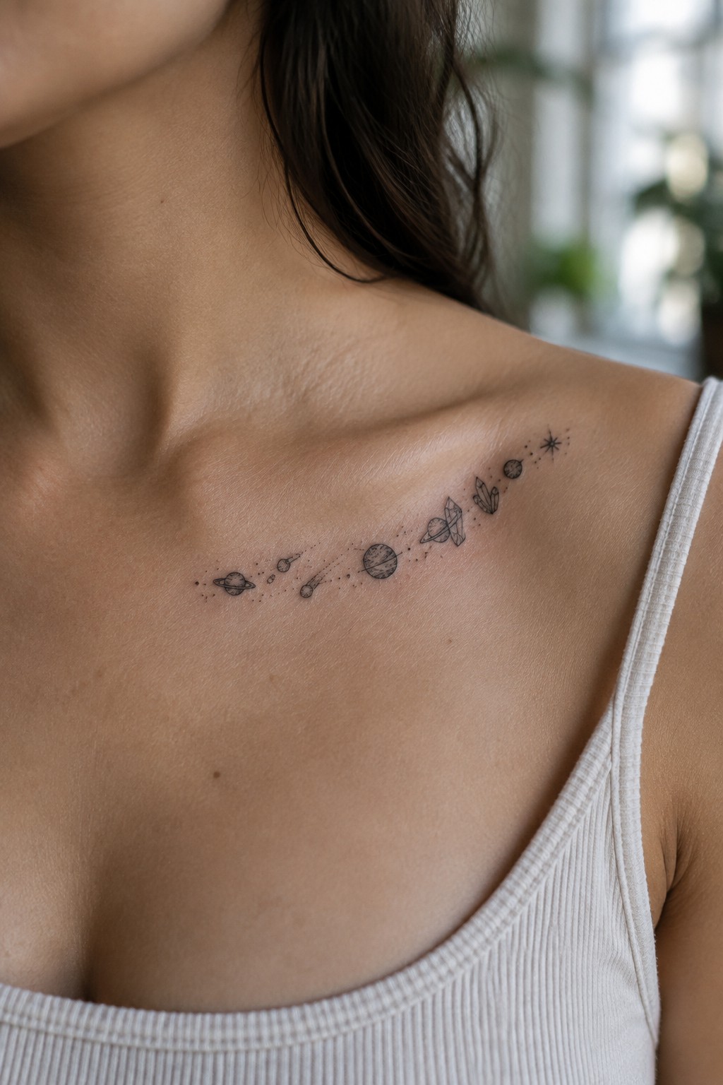 Woman's shoulder shows black celestial tattoo of planets, crystals, and stars above white tank top.