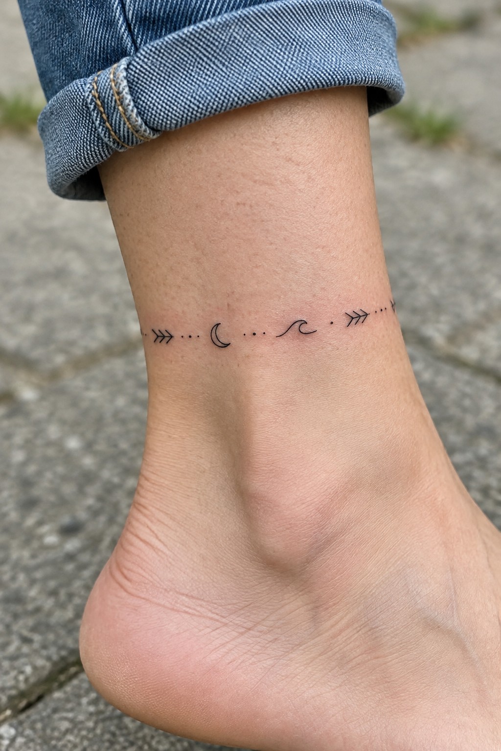 Bare ankle with moon, wave, arrows, and dots tattoo under rolled blue jeans.