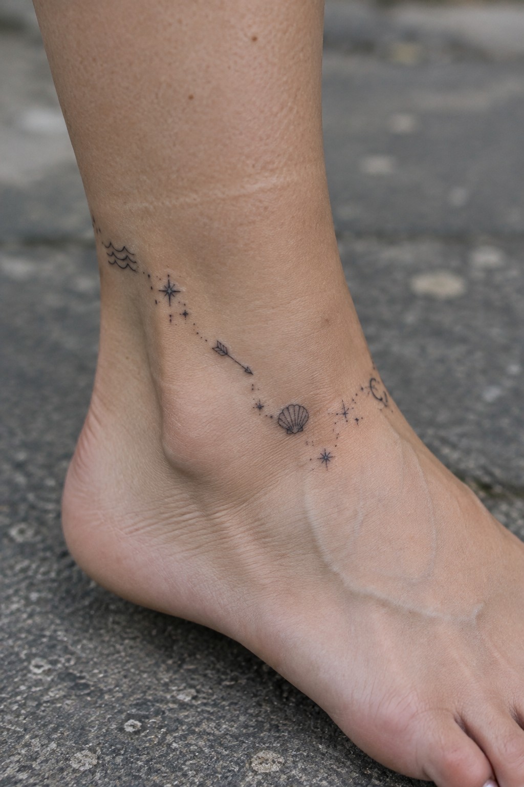 Bare ankle with celestial tattoos: waves, stars, arrow, seashell, moon on foot.