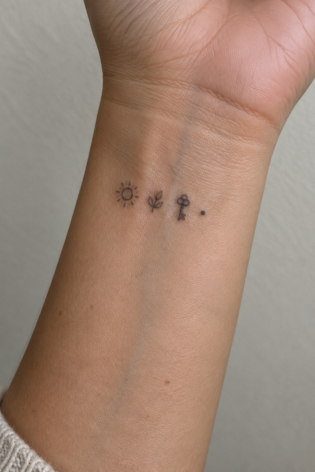 Light-skinned wrist with small sun, leaves, key, and dot tattoos above sleeve