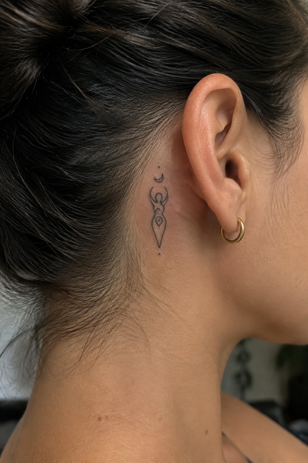 Dark hair in bun with crescent moon figure tattoo behind ear and gold hoop earring.