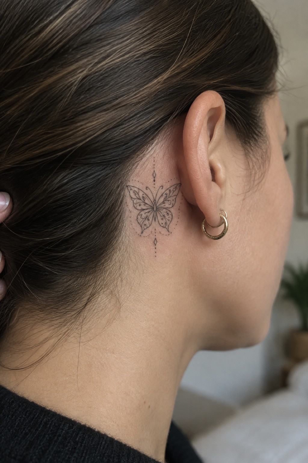 Brown-haired woman with ornate butterfly tattoo behind ear and gold hoop earring.