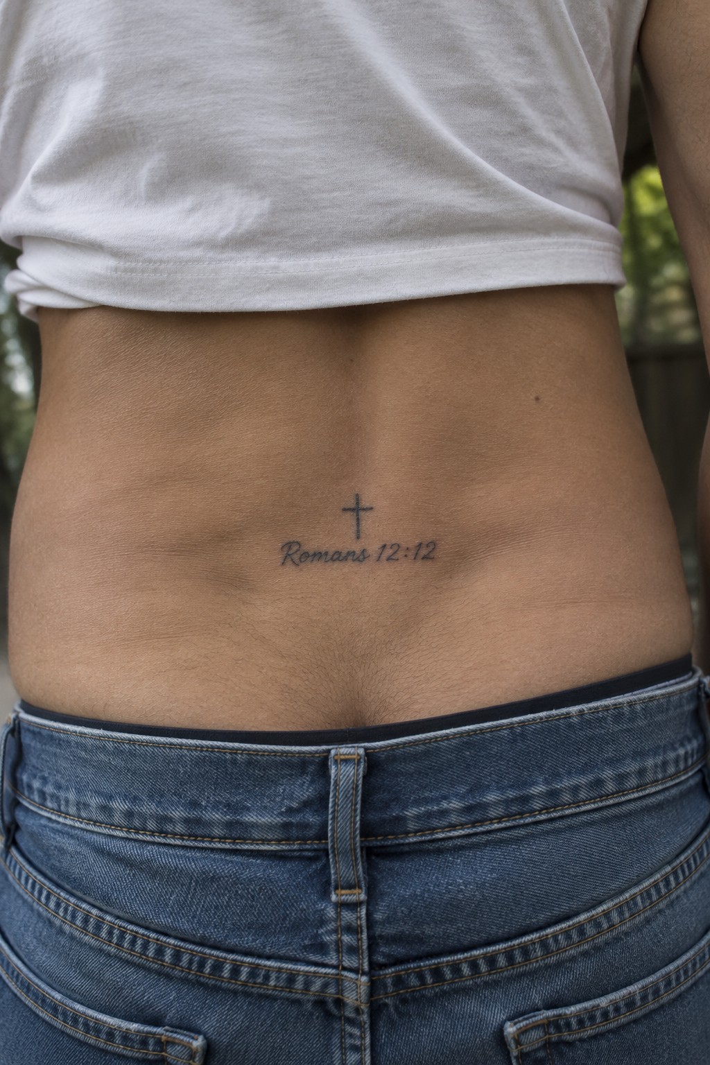 White t-shirt and blue jeans with cross tattoo reading Romans 12:12 on abdomen.