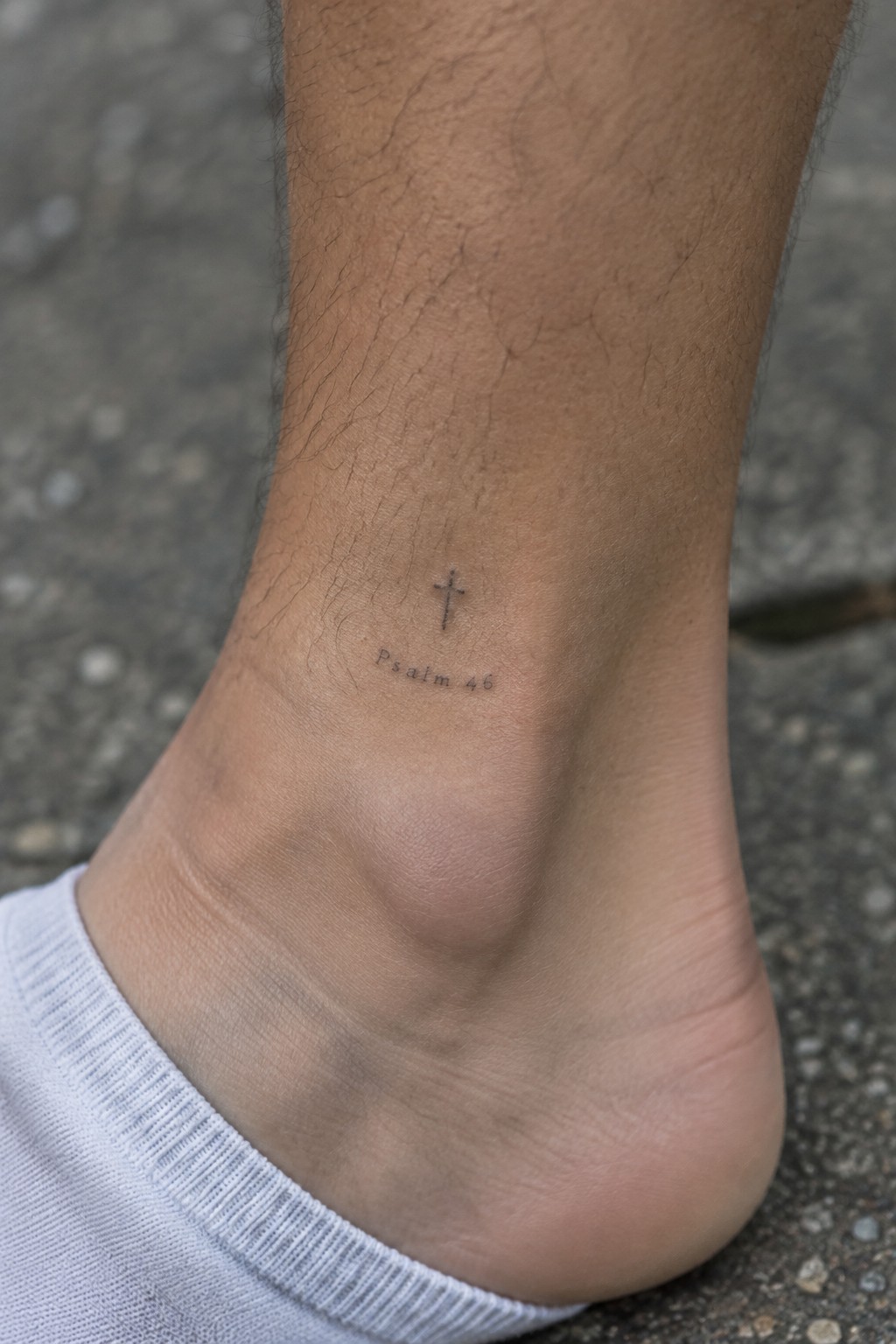 Cross and Psalm 46 tattoo on hairy ankle above white sock on pavement