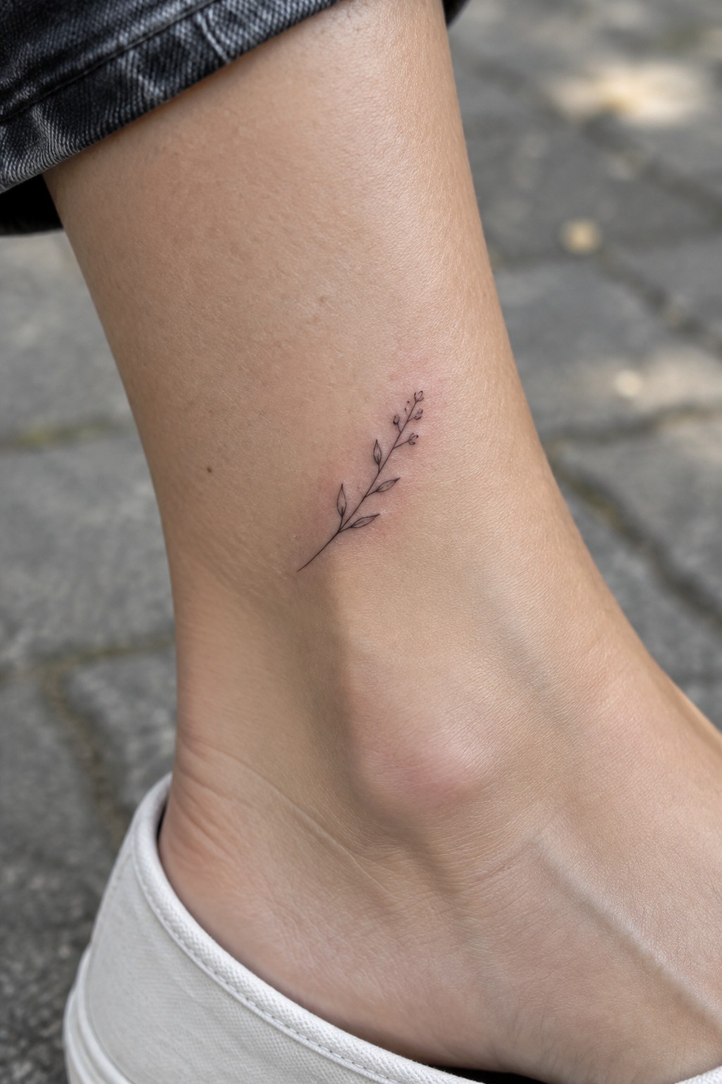 Small black leafy branch tattoo on ankle above white shoe on pavement.