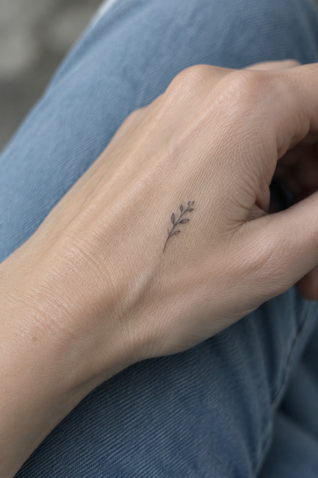 Small fine line botanical sprig tattoo on the back of a hand resting on blue jeans