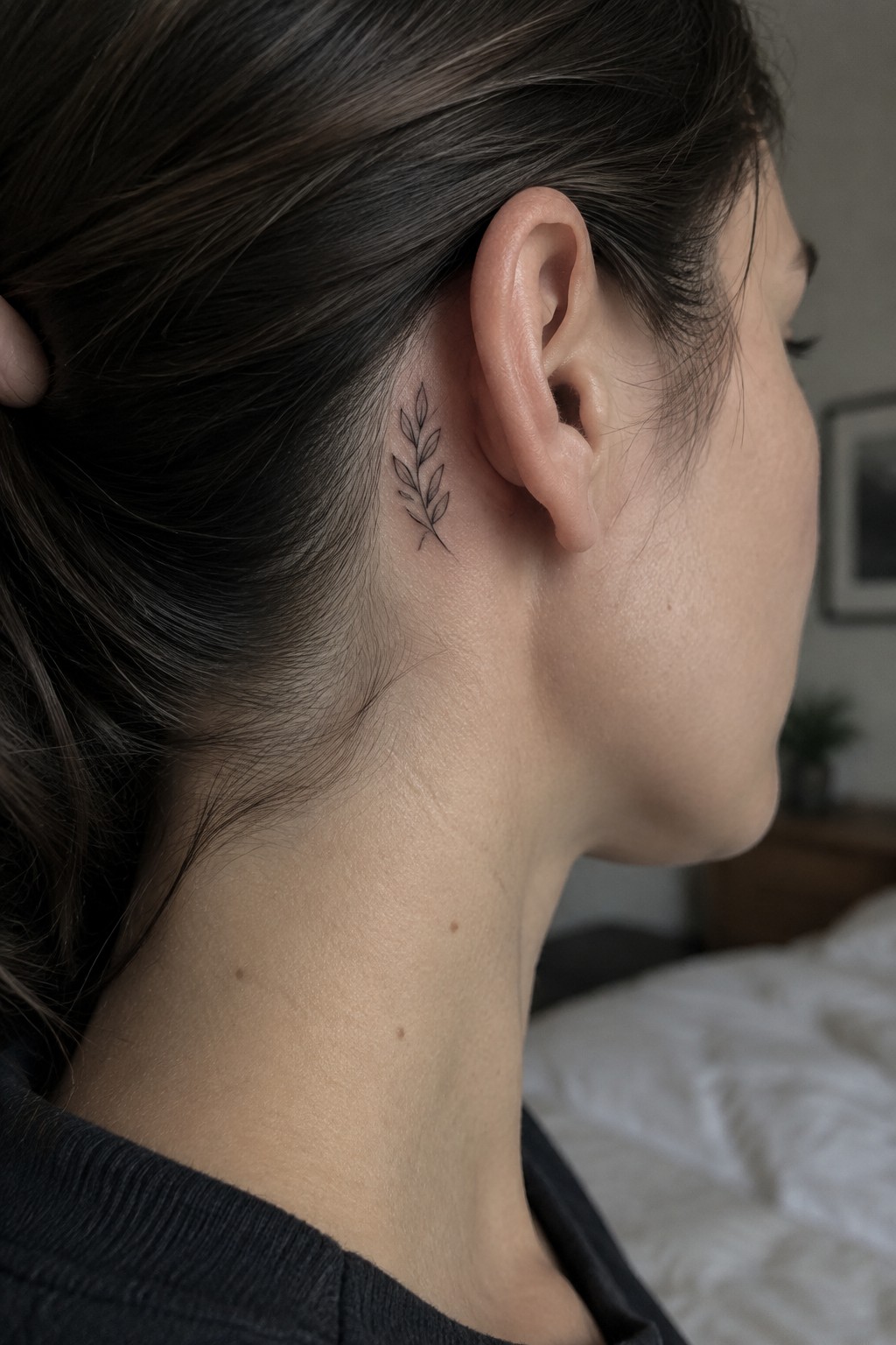 Woman with dark hair shows small leafy branch tattoo behind her ear.