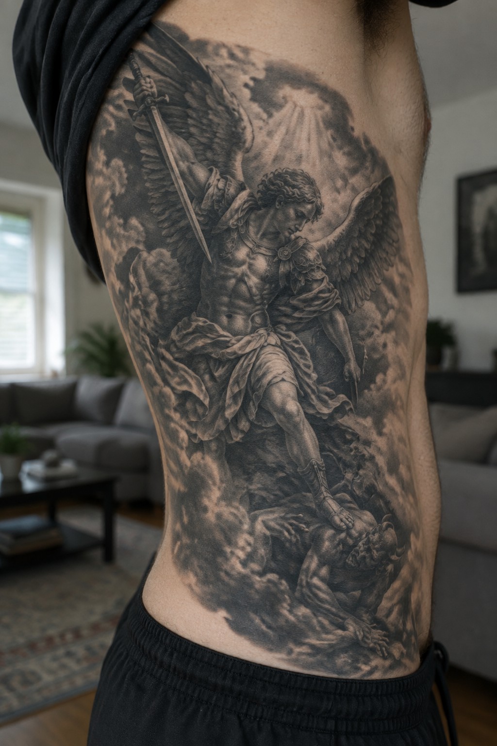 Large black-and-gray tattoo of winged angel with sword overpowering demon on torso