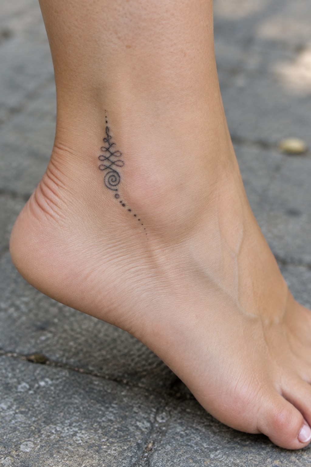 Delicate unalome tattoo with a spiral base and dotted line on the ankle.