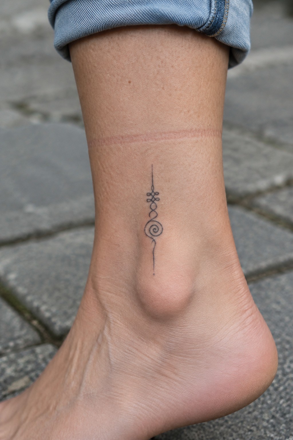 Ankle with black spiral tattoo under rolled blue jeans on stone pavement