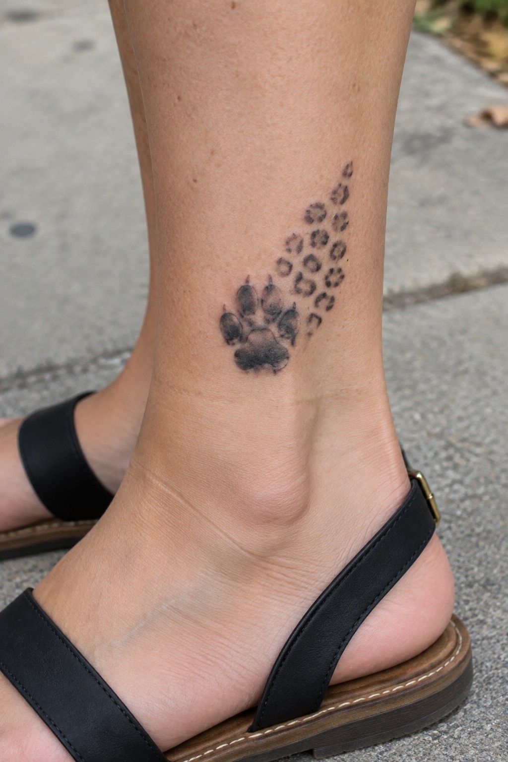 Black paw print tattoo with smaller prints trailing up ankle above black sandal