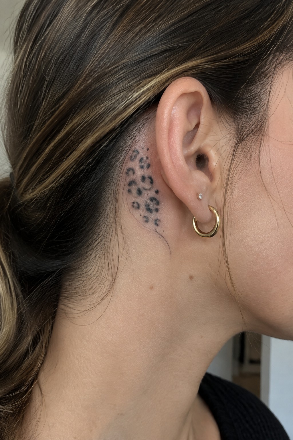 Leopard-print tattoo behind ear with gold hoop and diamond stud earrings in highlighted hair.