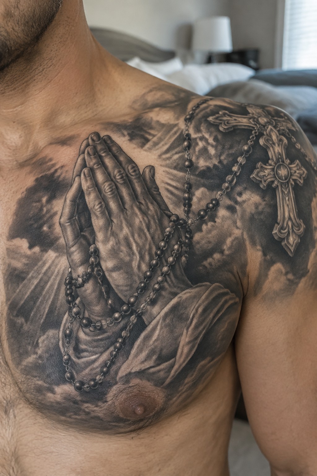Realistic black and gray tattoo of praying hands holding a rosary on a man's chest with a cross on the shoulder and soft cloud shading in the background
