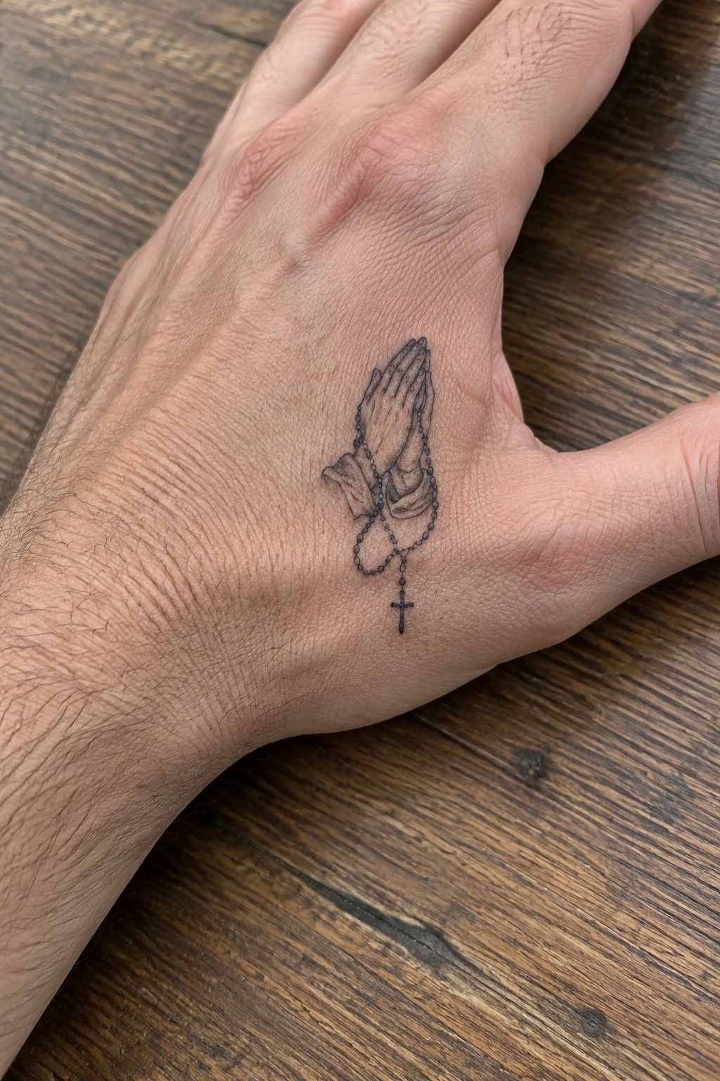 Hand on wooden table with black praying hands and rosary tattoo