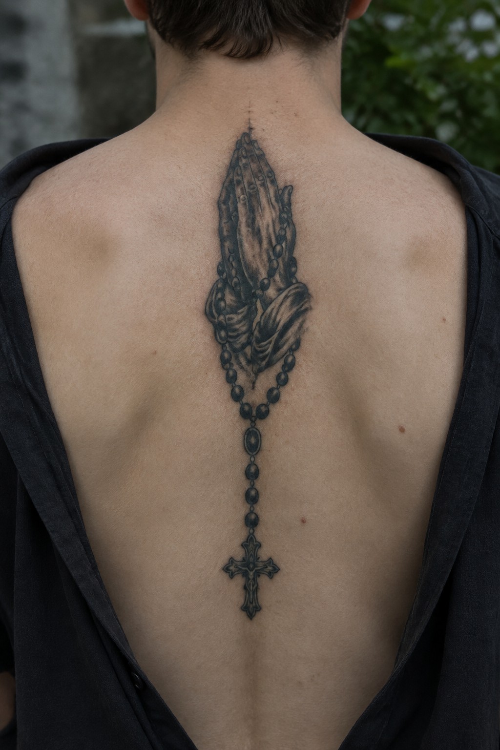 Tattoo of praying hands holding rosary beads with cross on person's bare back