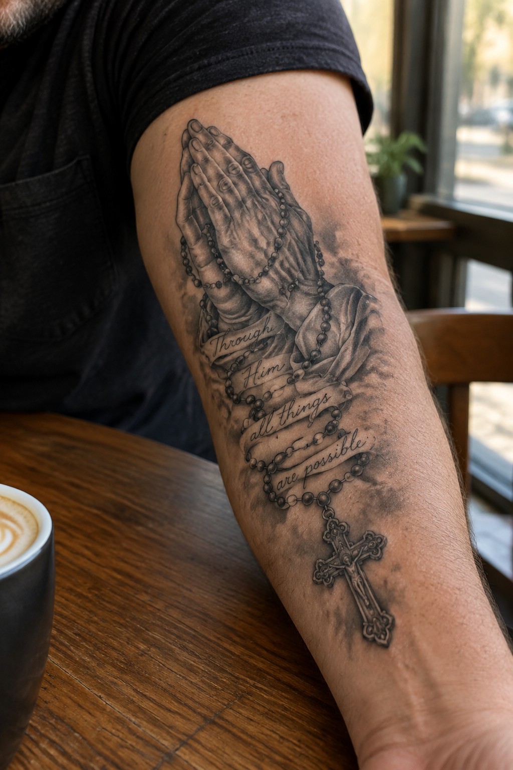Black and grey realistic tattoo of praying hands holding rosary beads with a banner quote and a hanging cross on a man's forearm.