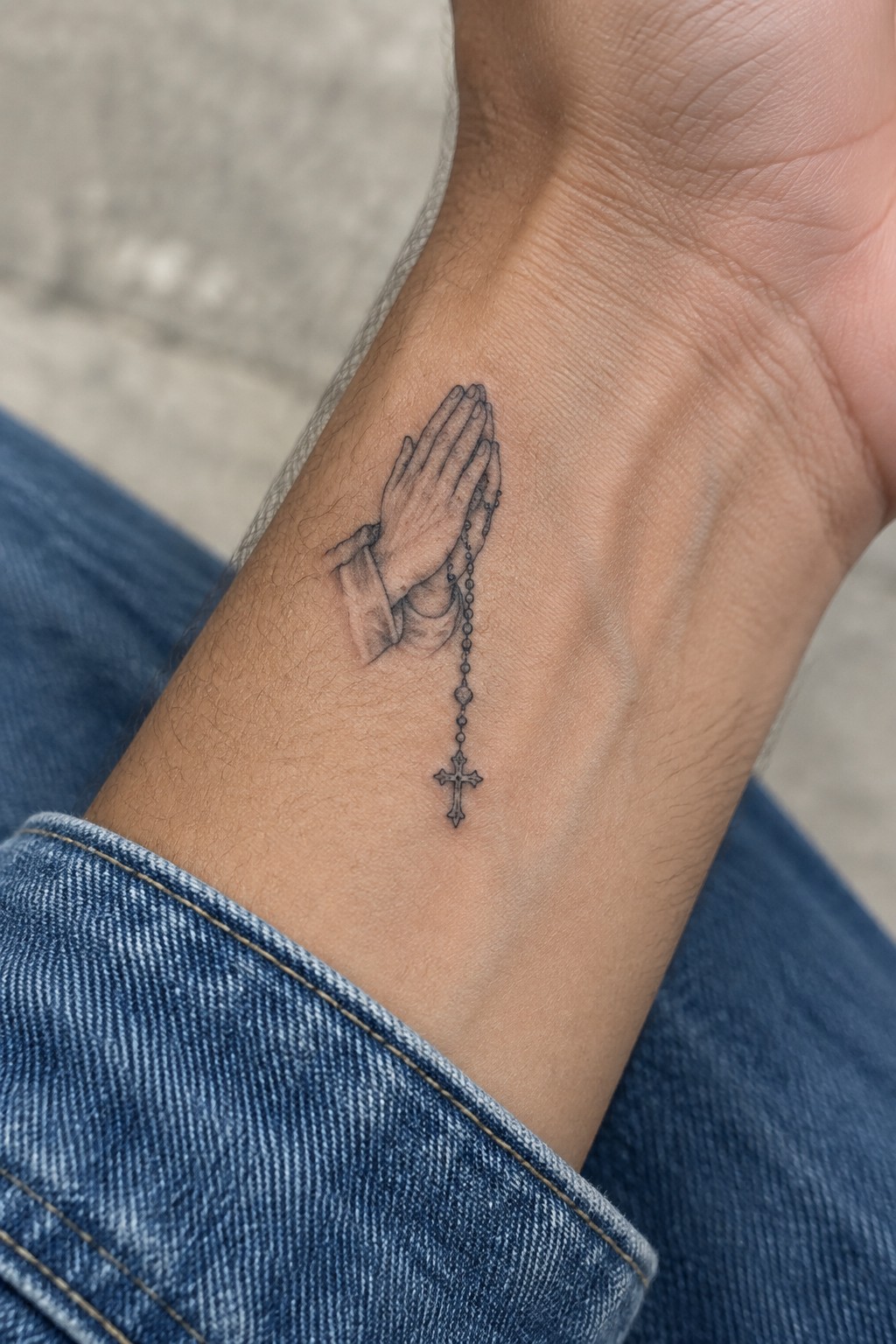 Praying hands rosary cross tattoo on wrist above blue denim jeans.