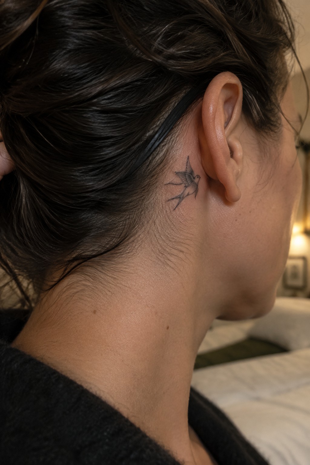 Small bird tattoo behind ear of person with dark hair and black sweater